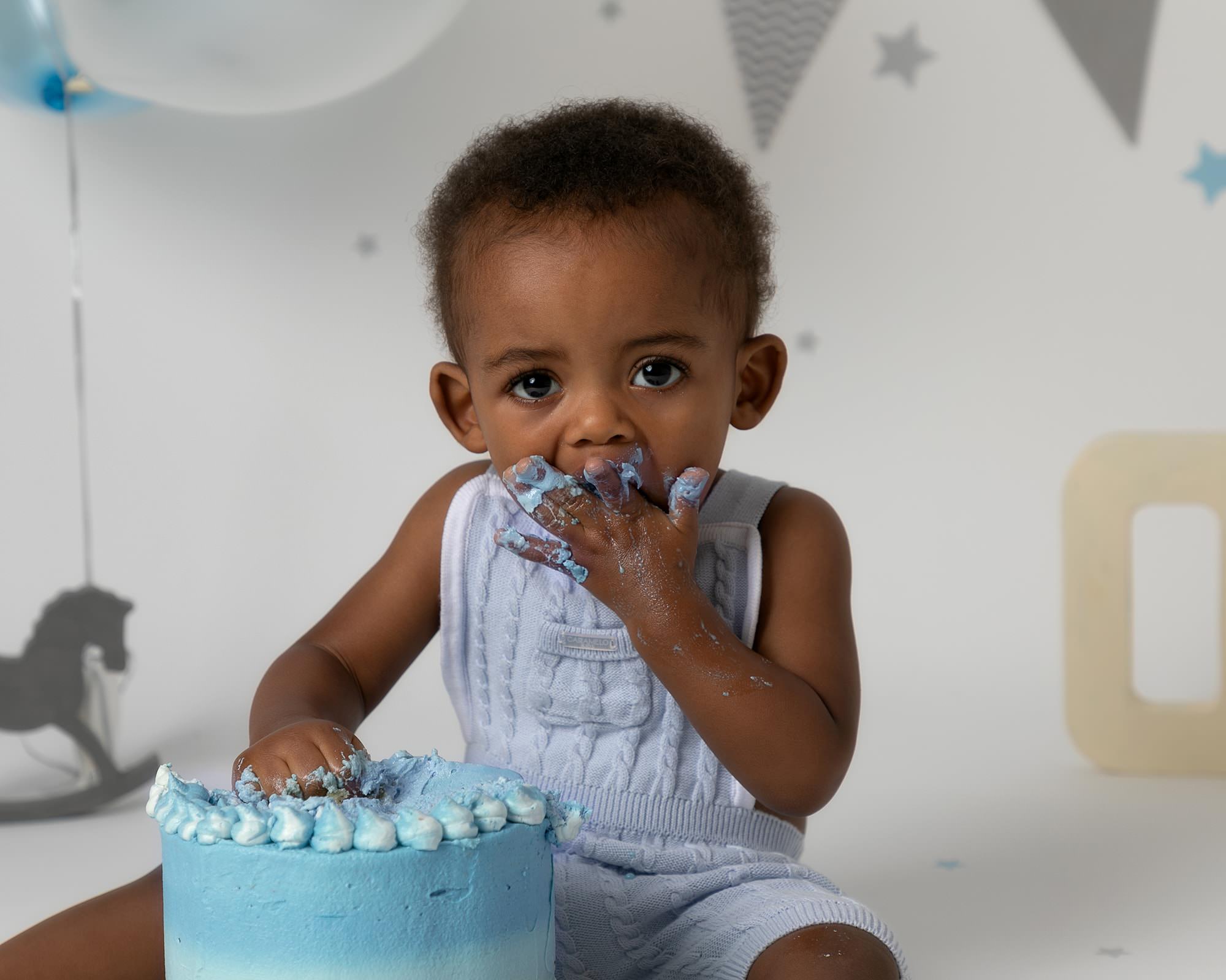 Baby boy in blue romper. Close up image of his face as he uses his hand to eat cake with blue icing at his 1st birthday cakesmash photoshoot in Glasgow