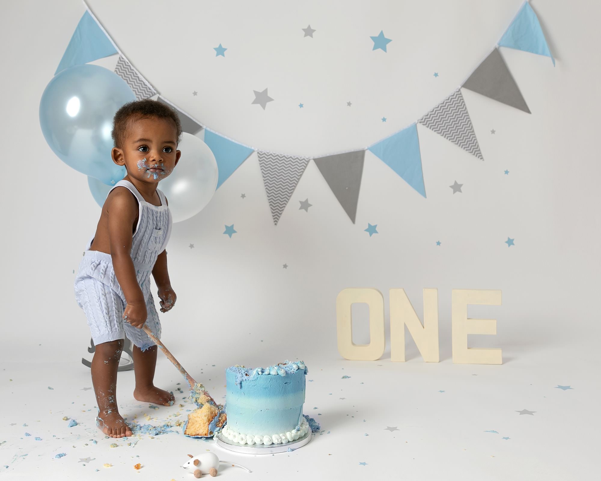 Baby boy in a blue romper, smiling as he touches his birthday cake made of blue buttercream. Baby is at his 1st birthday photoshoot in Glasgow sat on a white backdrop with blue & grey bunting, balloons and stars