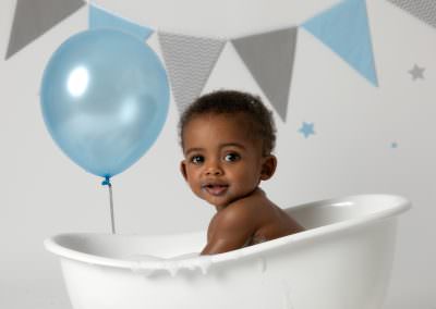 Baby boy, looking at the camera, sat in a white bath with a blue balloon and grey & blue bunting in the background. Image taken at his 1st birthday cakesmash photoshoot in Glasgow