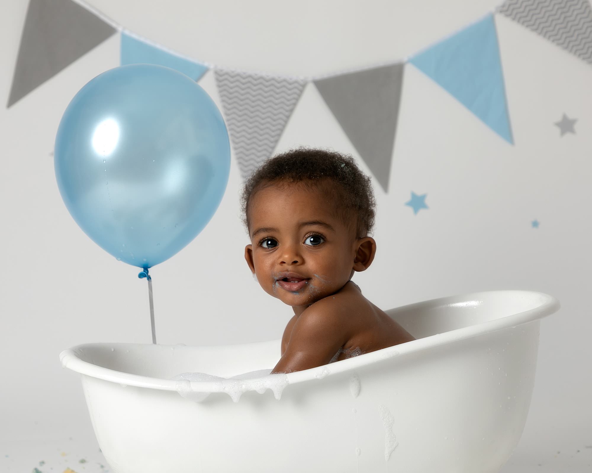 Baby boy, looking at the camera, sat in a white bath with a blue balloon and grey & blue bunting in the background. Image taken at his 1st birthday cakesmash photoshoot in Glasgow