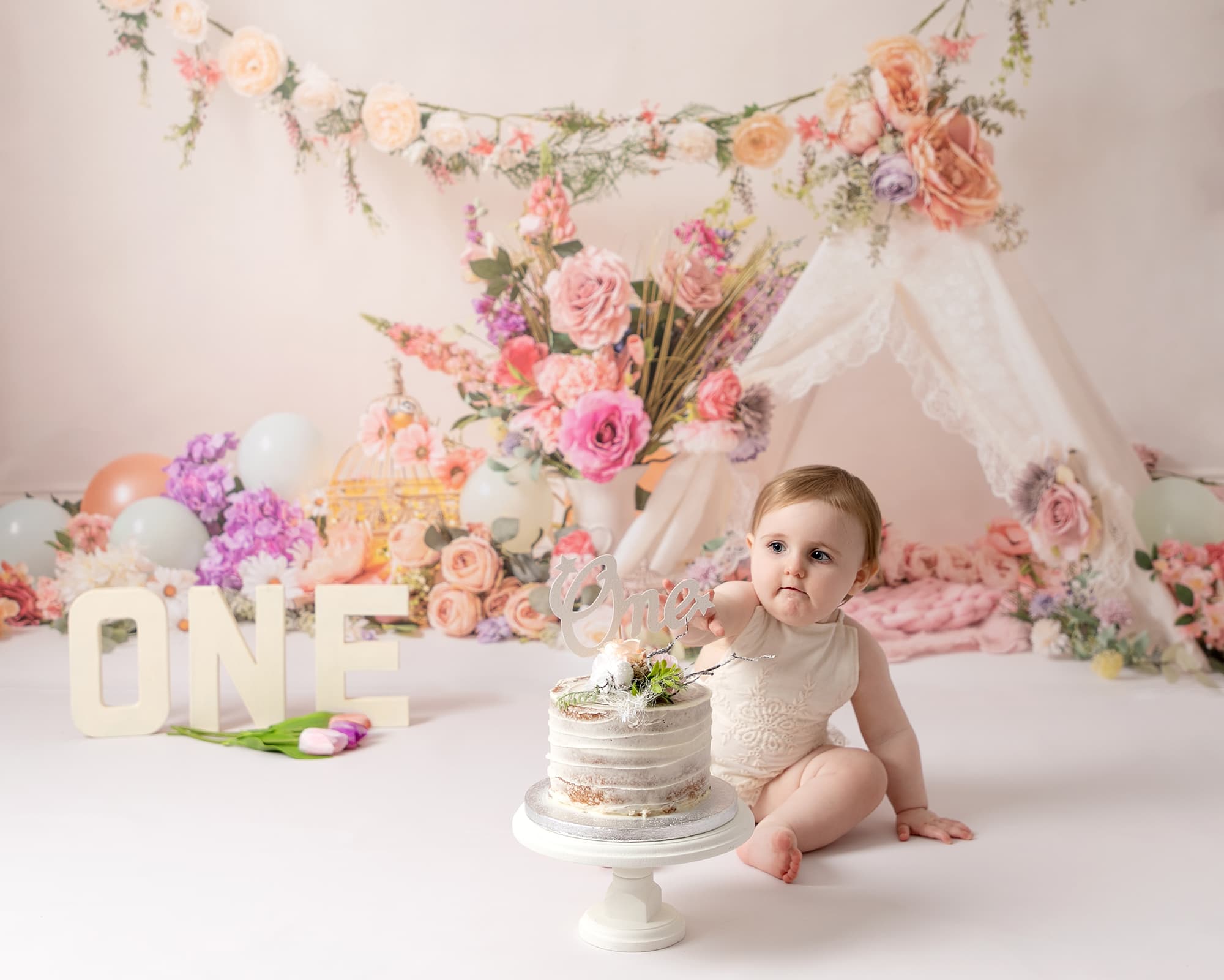 Baby girl wearing a cream romper sat looking at her birthday cake. Baby is at her 1st birthday photoshoot in Glasgow. Backdrop of floral arrangement and a teepee