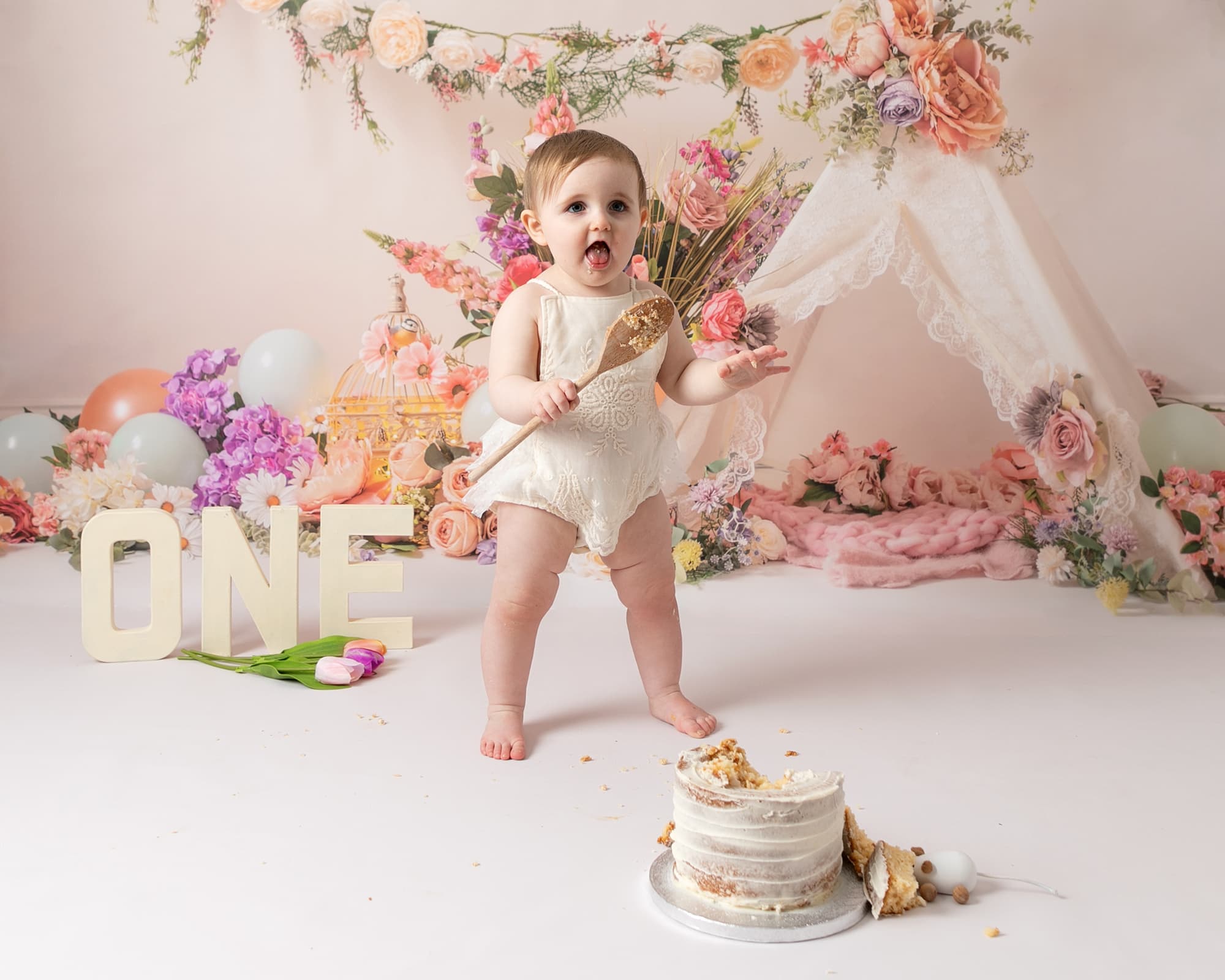 baby girl standing up holding a wooden spoon, wearing a cream frilly romper. Baby is at her 1st birthday photoshoot in Glasgow. Floral background in pink & peach tones with a teepee