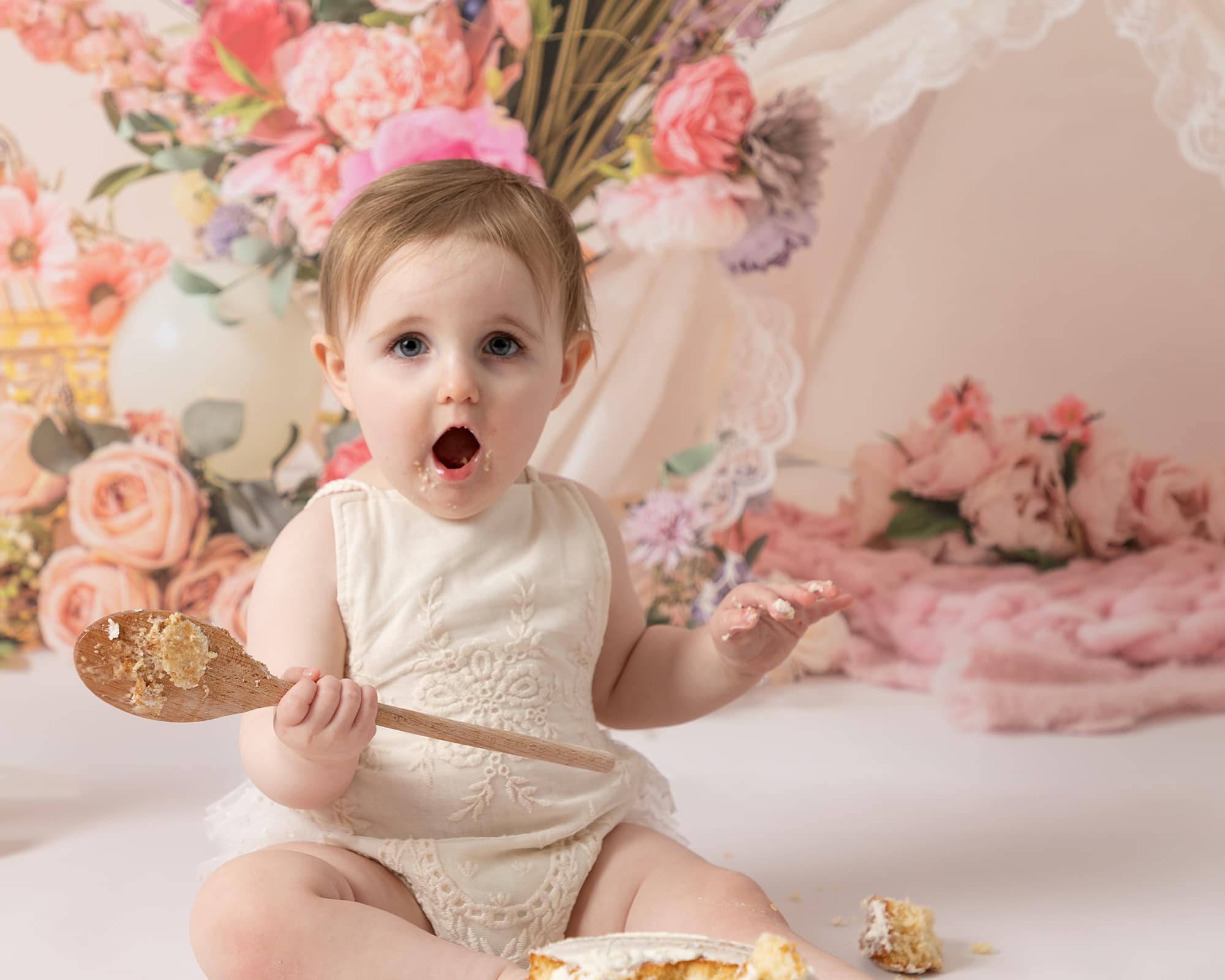 Baby girl wearing a cream romper sat looking at her birthday cake. Baby is at her Cakesmash Birthday Photoshoot in Glasgow. Backdrop of floral arrangement and a teepee