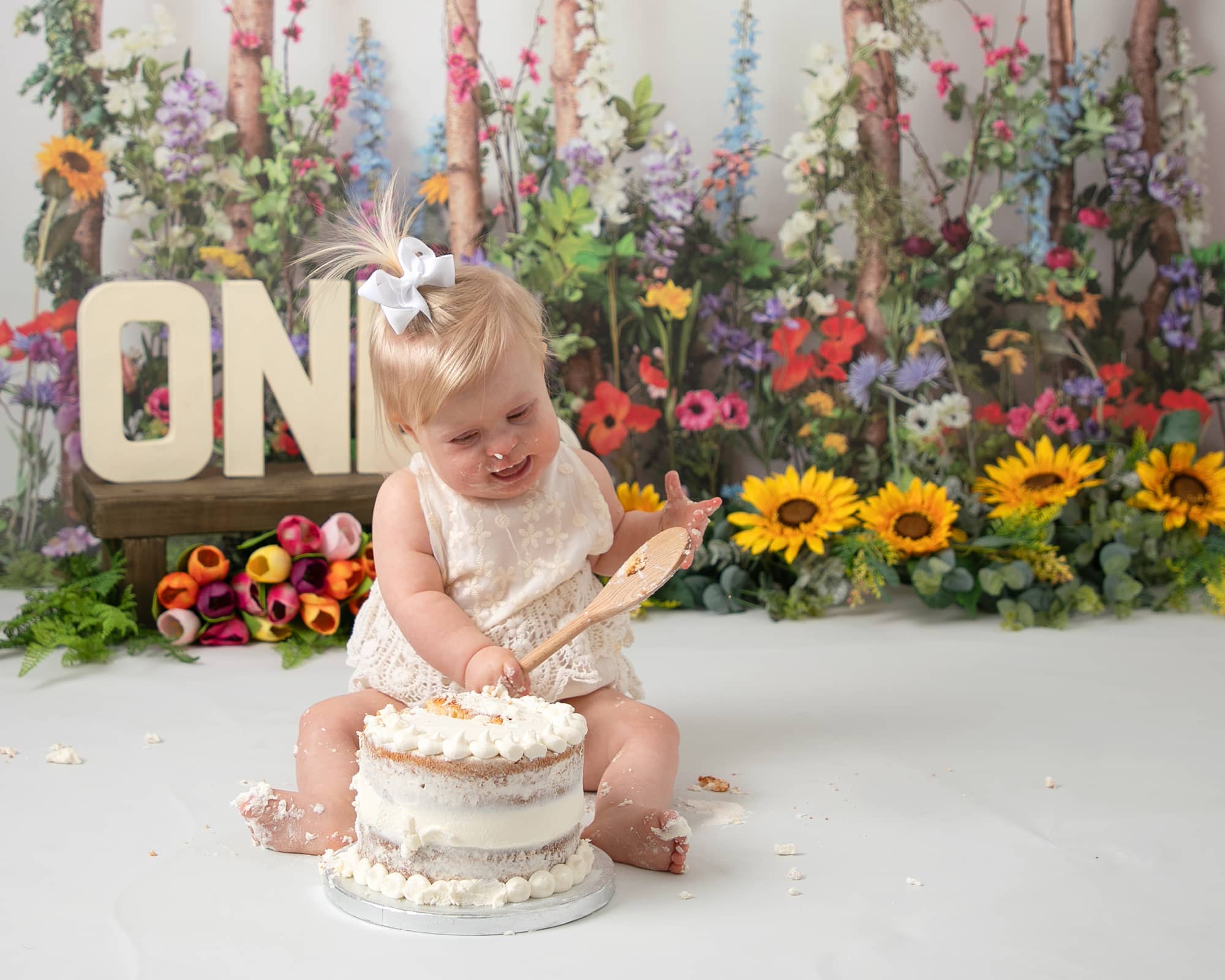Baby girl sat behind a cake playing with a wooden spoon. using a meadow style backdrop with lots of flowers