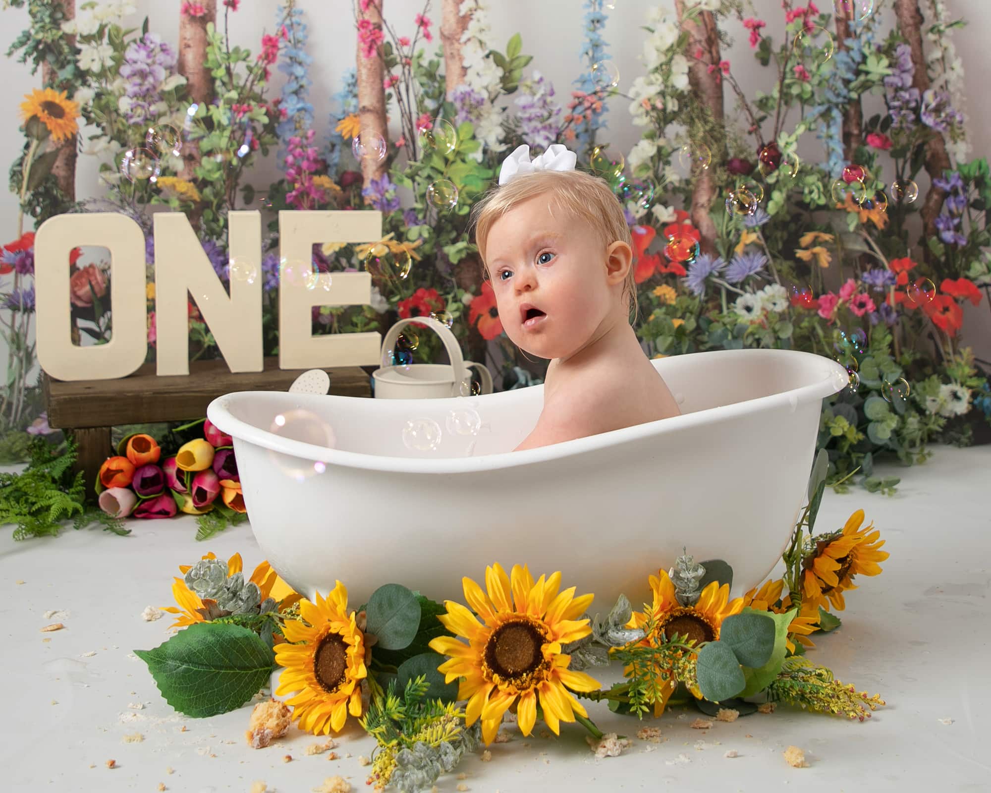 Baby girl sat in mini white bathtub, surrounded by sunflowers. using a meadow style backdrop with lots of flowers