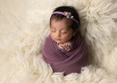 Baby girl sleeping, wrapped in purple with purple floral headband on a cream flokatti. Image part of newborn photography portfolio in Glasgow