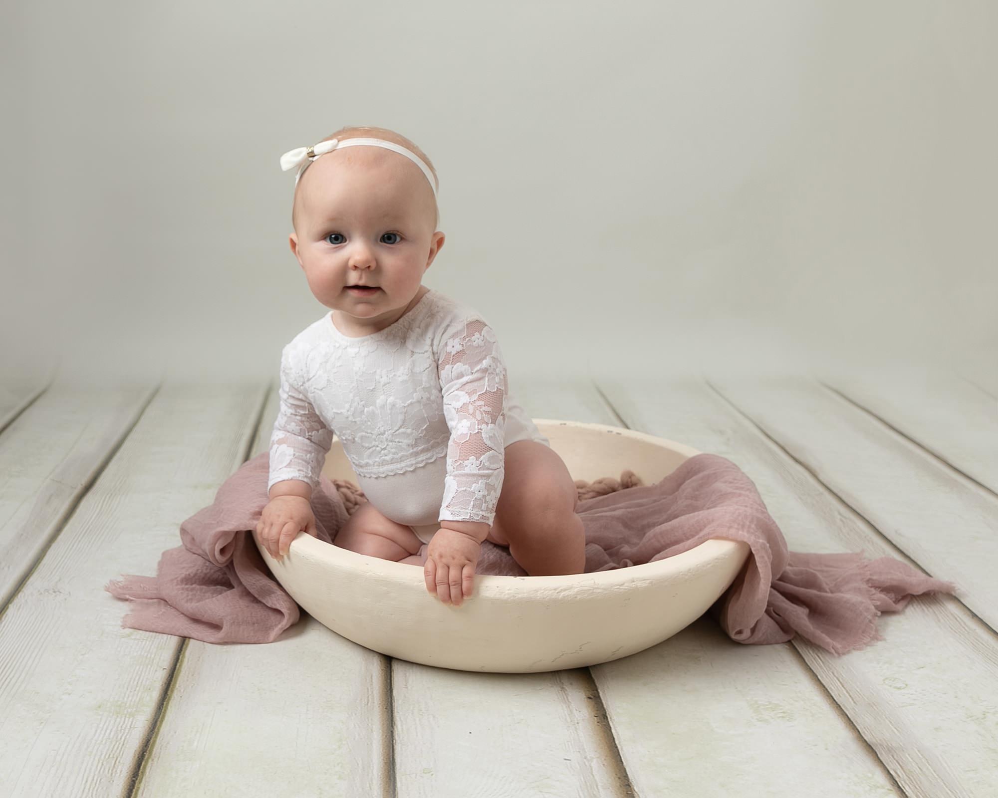 Baby is on a cream backdrop at her photoshoot with Baby photographer in Glasgow. Sat in a cream bowl wearing a white romper