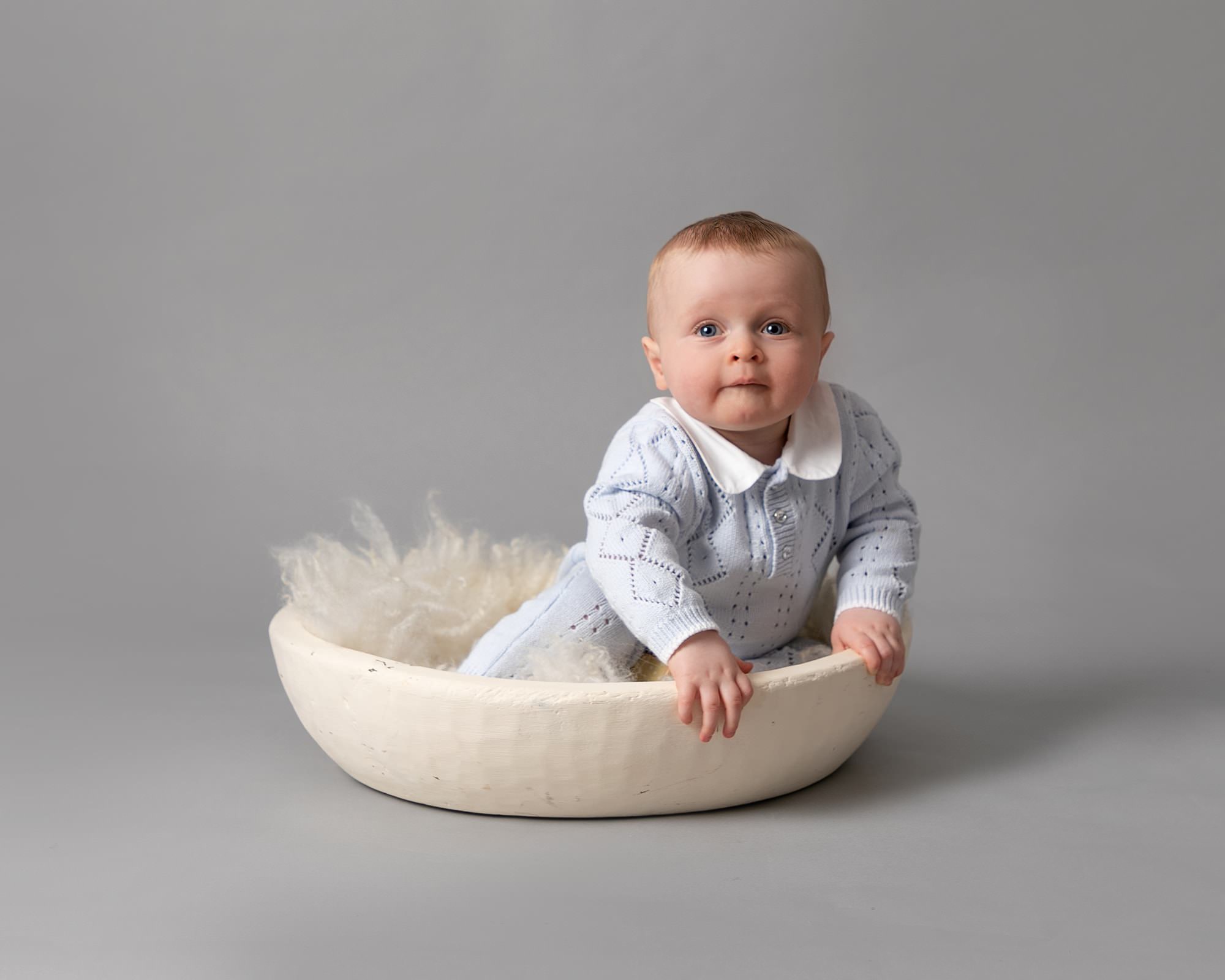 Baby boy on grey backdrop, sat in a cream bowl with fluff. Blue romper