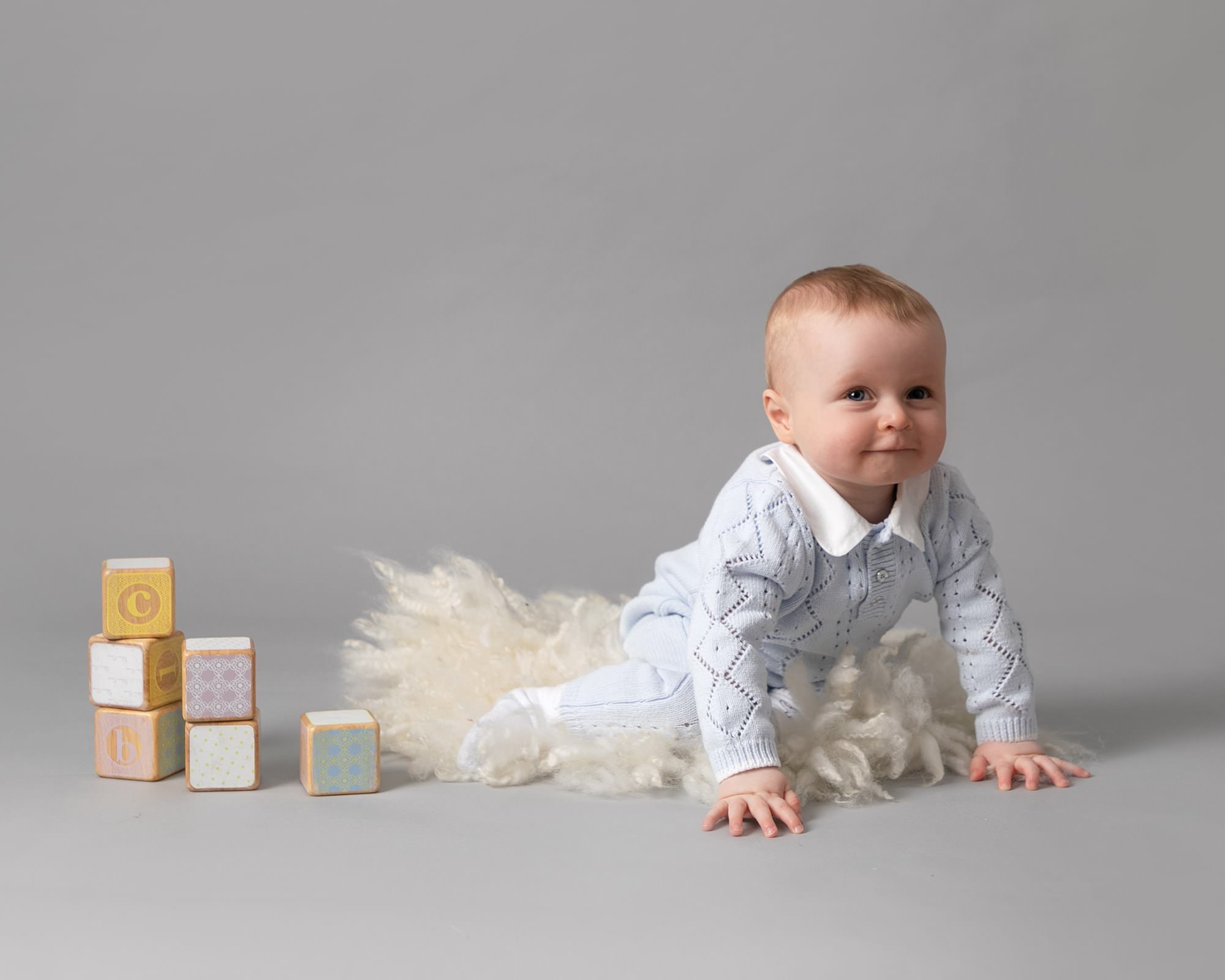 Baby boy on grey backdrop, sat in a cream bowl with fluff. Blue romper
