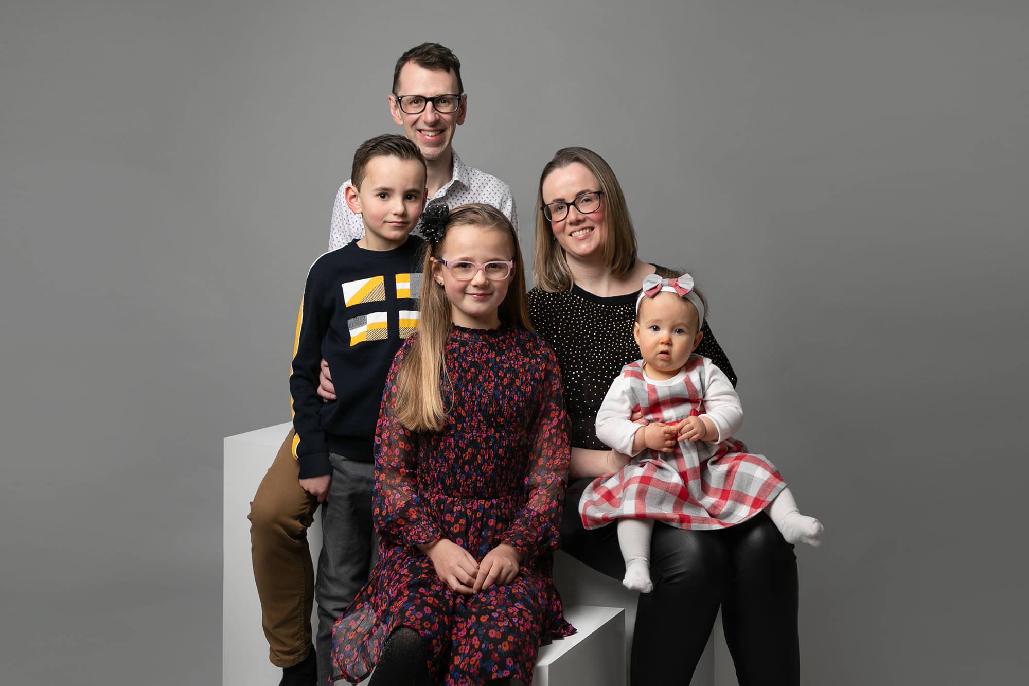 Family of 5 during photoshoot in Glasgow. Image taken on grey backdrop. Girl age 8 wears black dress with floral print. sibling baby sits on Mum's lap wearing red grey & white check dress. Boy sits on Dad's lap wearing black T with yellow pattern. Image by Baby Photographer in Newton Mearns