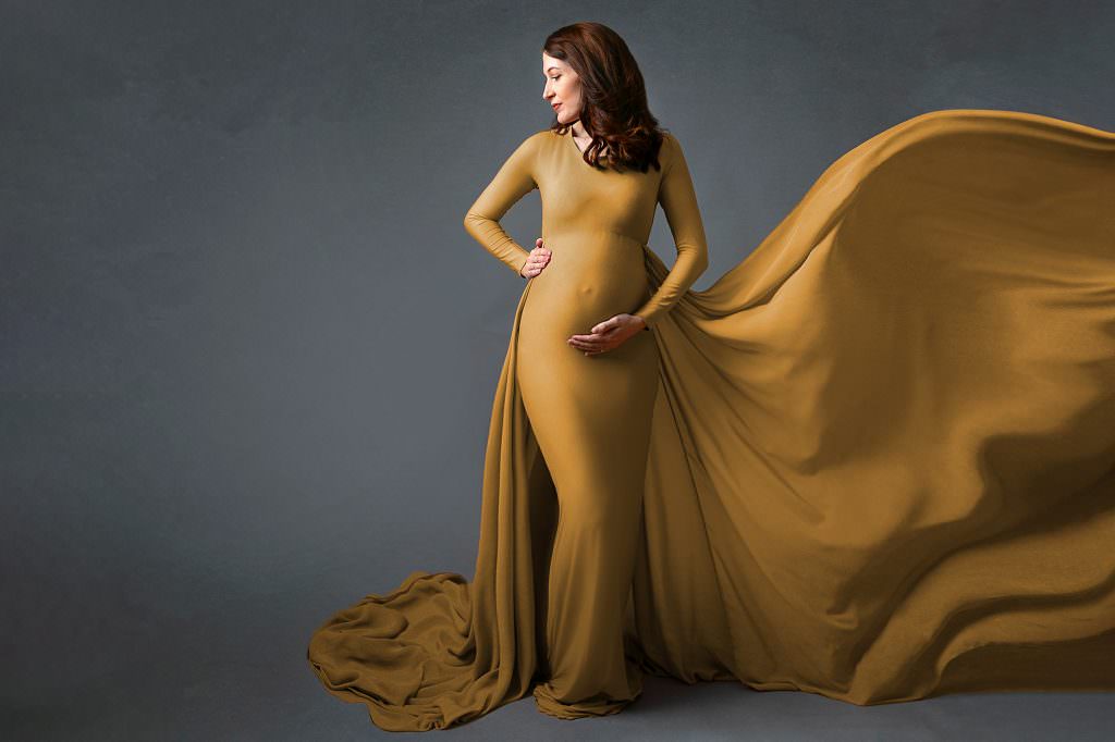 Pregnant lady with red hair wearing an ochre tight fitted gown. Female looks over her left shoulder and fabric flows in the air to her right. Image taken by Glasgow Baby & Maternity Photographer