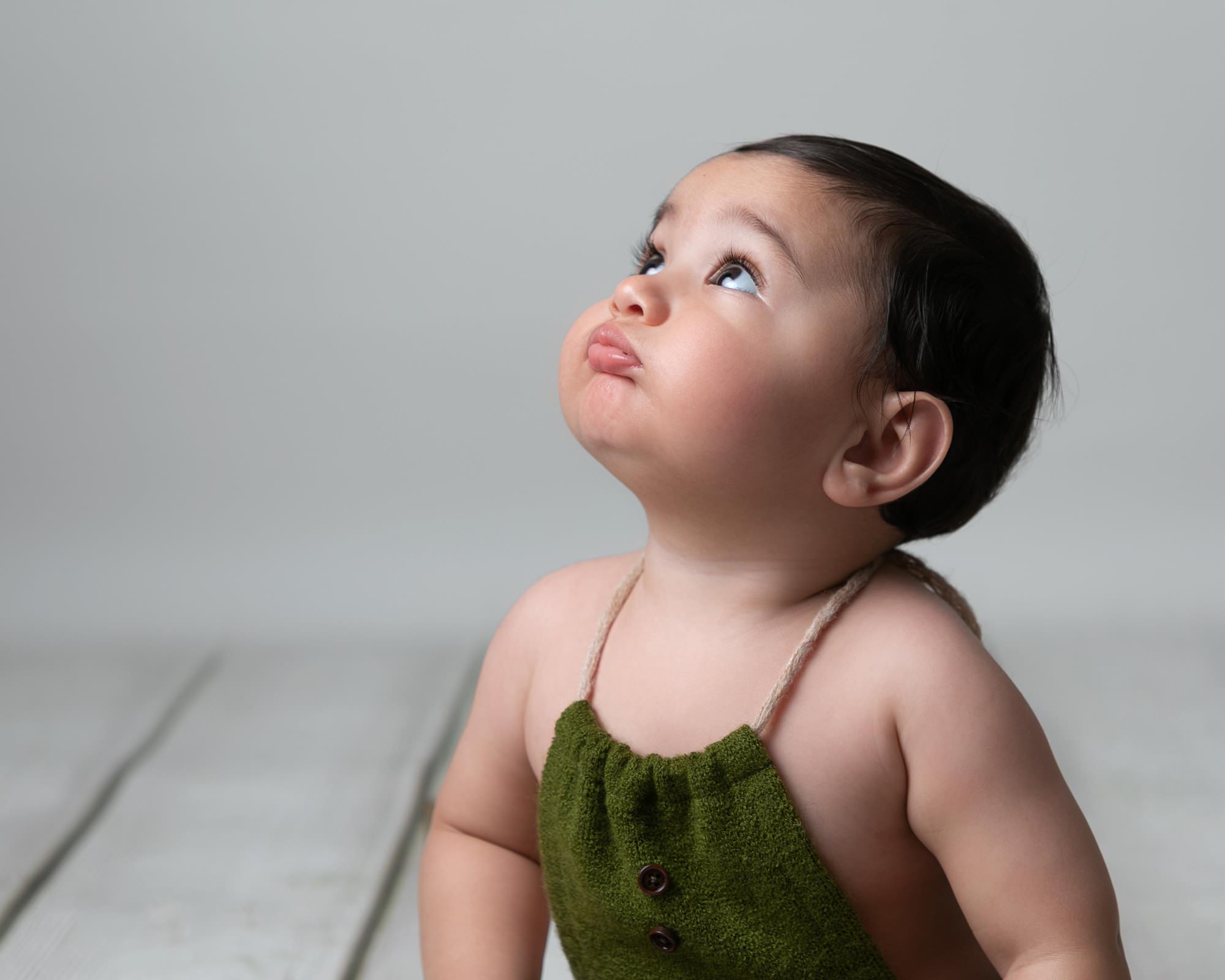 Baby boy in green dungarees looking upwards and blowing a raspberry. Image taken at photoshoot with Glasgow Baby Photographer