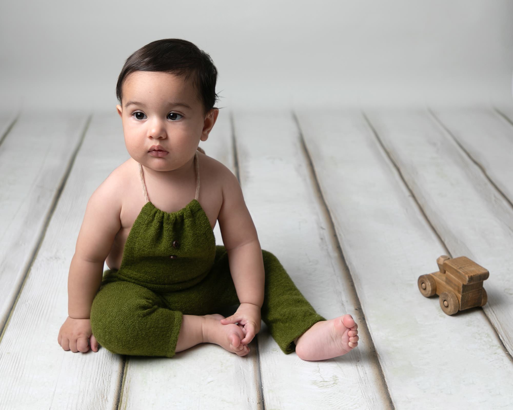 Baby boy in green dungarees, sat on cream wooden floor with wooden toy car. Image taken at photoshoot with Glasgow Baby Photographer