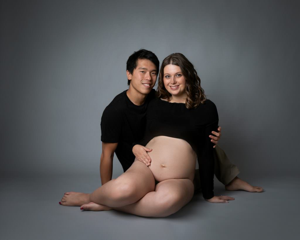 Pregnant female and male partner sat on the floor of a grey backdrop. Female wears cropped black polo neck and make wears beige chinos and black short. Image taken at Maternity photography session in glasgow