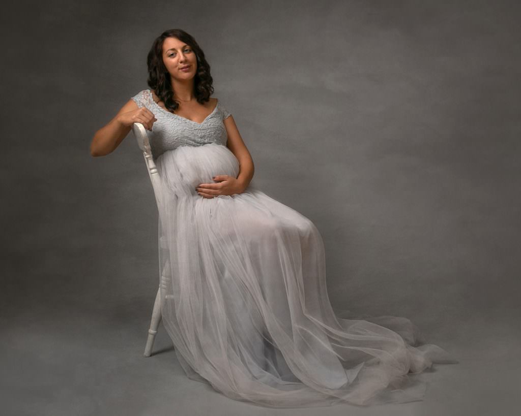 Female wearing maternity gown in grey with tulle skirt, sitting on a white wooden chair on a grey backdrop. Maternity photoshoot Glasgow