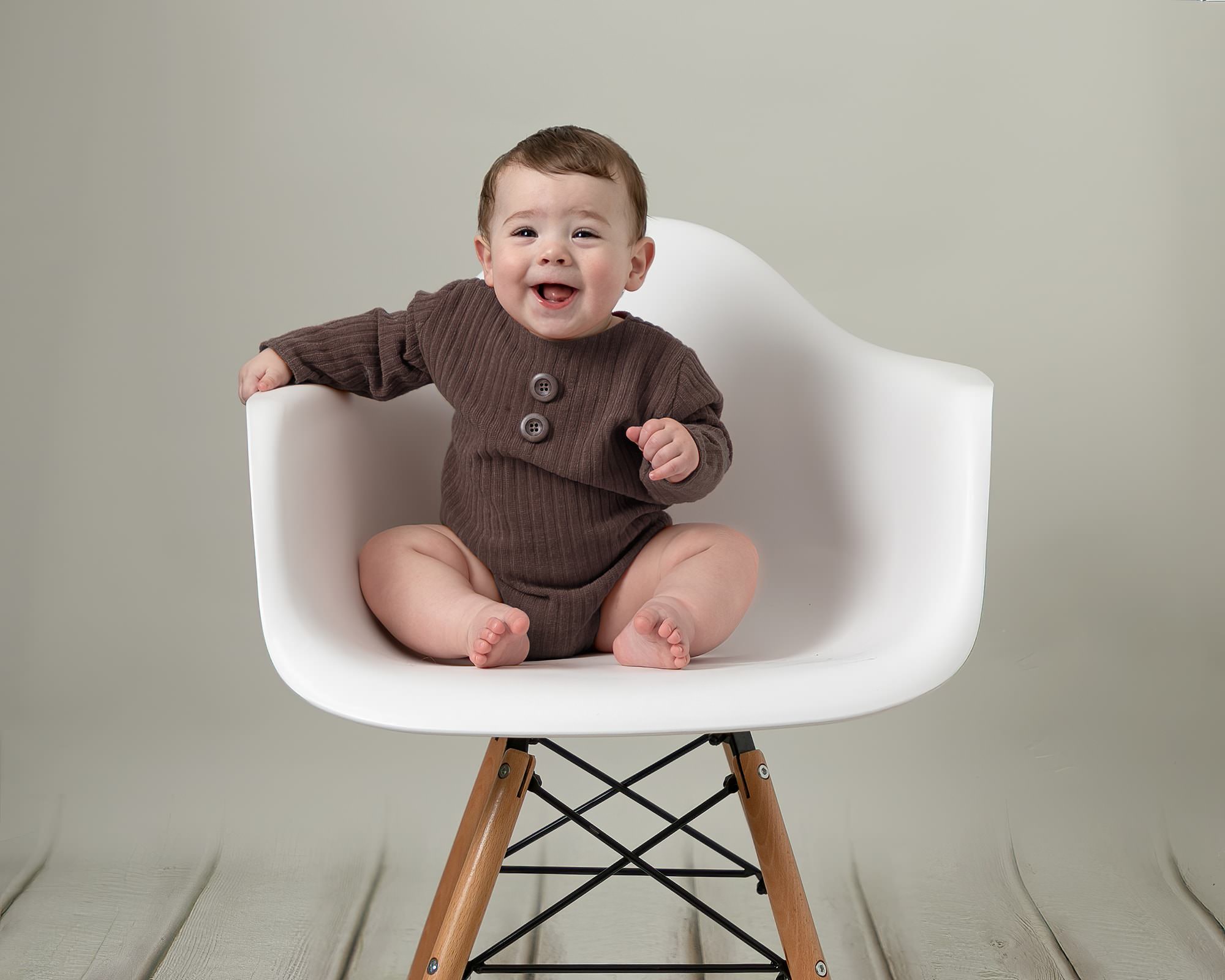 Baby boy sat on a white chair laughing. Boy is wearing a brown romper. Part of a gallery by Glasgow Baby Photographer
