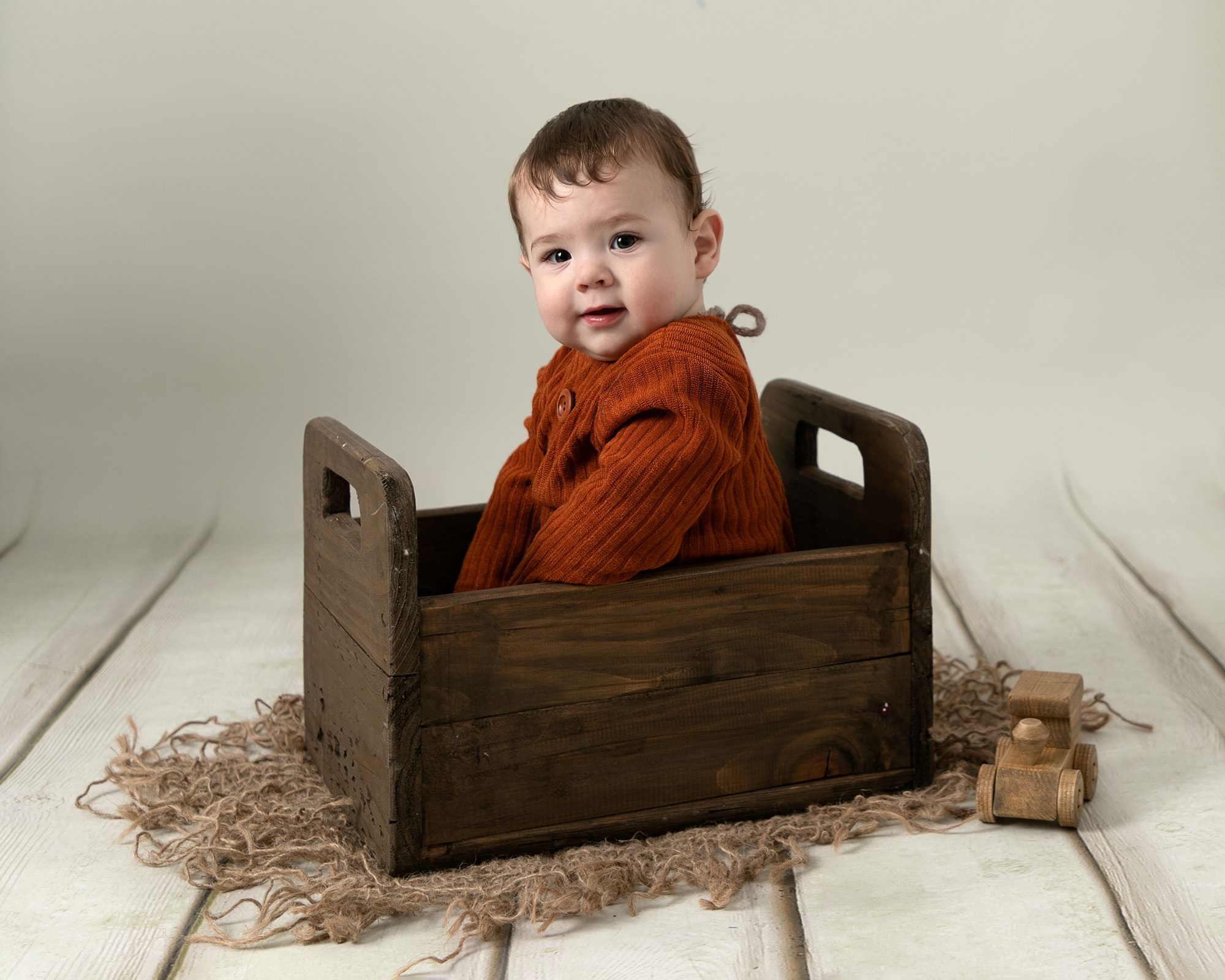 Baby boy wearing a rust romper sat in a brown crate on a cream backdrop.