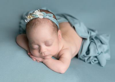 Baby mil laying on her tummy, chin on hands. baby posed by Glasgow baby photographer during newborn photoshoot. Baby lies on a duck egg blue backdrop with a floral tie back and matching blanket over her back