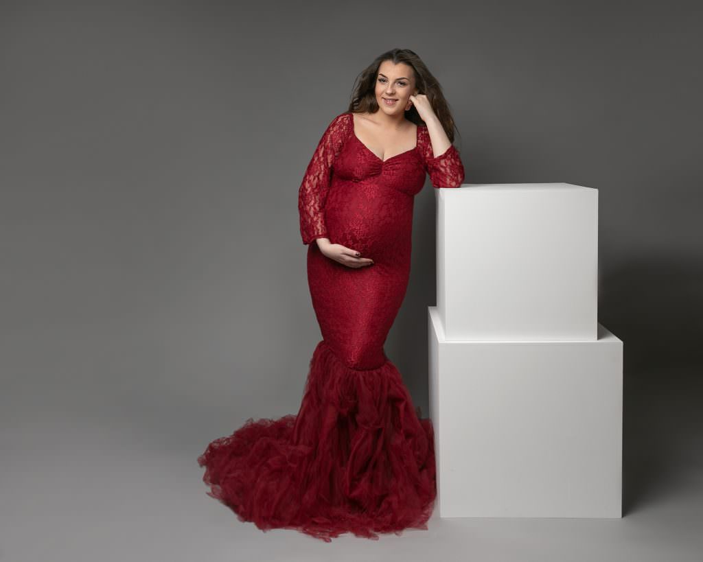 Female wearing a red fishtail, lace gown stands leaning against white blocks. Posing for her pregnancy photoshoot. Subject is smiling and holding her bump. Image taken by Glasgow photographer