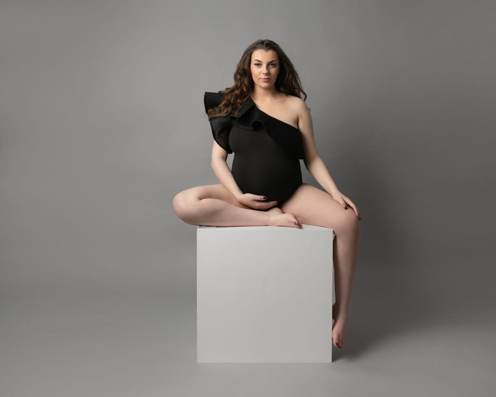 pregnant woman with long dark hair, wearing a black body suit, sat facing the camera on a white posing block. Image taken maternity photography session in Glasgow