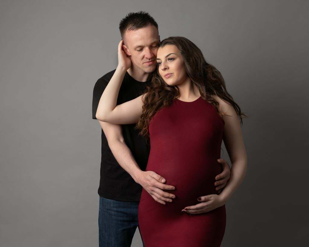 Male & female with arms around each other. Female wears a red maternity dress and male black t shire & jeans. Couple are posed during their pregnancy photoshoot taken at a studio in Newton Mearns Glasgow