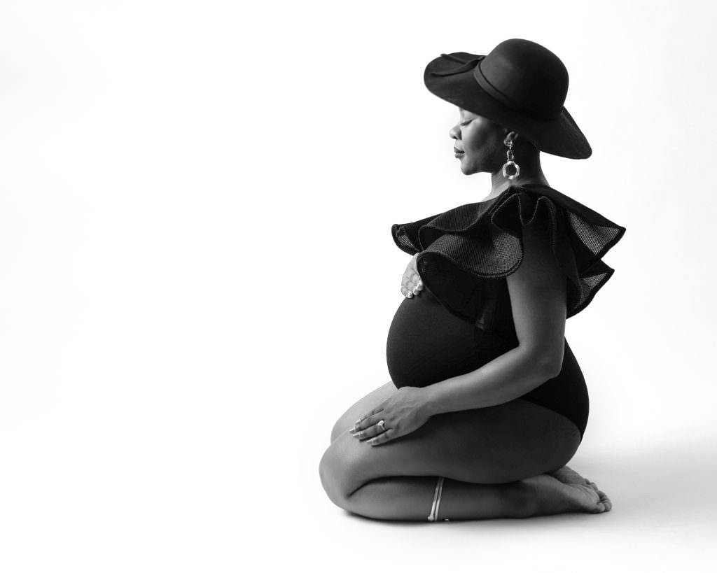 Pregnant female sat on her knees on a white backdrop, posing for her pregnancy photoshoot. Female wears a black bodysuit and black hat. Taken at Glasgow photography studio