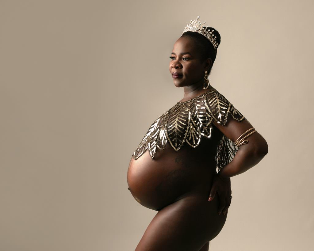 Female wearing her hair in a bun and a sparkly cape covering her chest. Heavily pregnant with naked bump, standing in profile view on a cream backdrop. Image is part of a pregnancy photoshoot taken in Glasgow