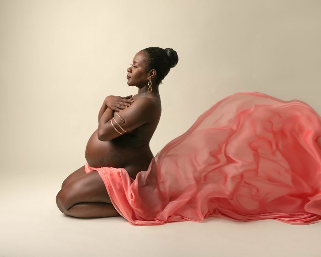 Female kneeling on the floor with peach fabric draped over her lap and behind. Subject is covering her chest with her arms and looking forward. Images part of a pregnancy photoshoot in Glasgow scotland