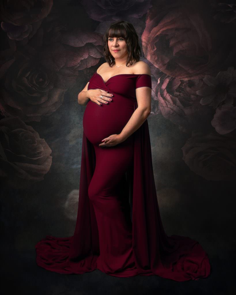 Female wearing red gown, dark hair holding her pregnancy bump. Floral backdrop used during her photoshoot in Glasgow
