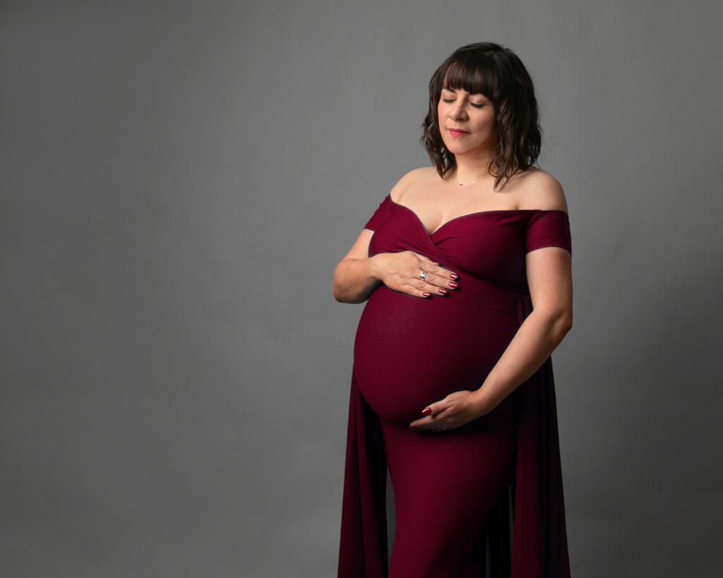 Pregnant lady wearing a red gown at her pregnancy photoshoot in Glasgow. Female stands with her eyes closed, hands on her bump