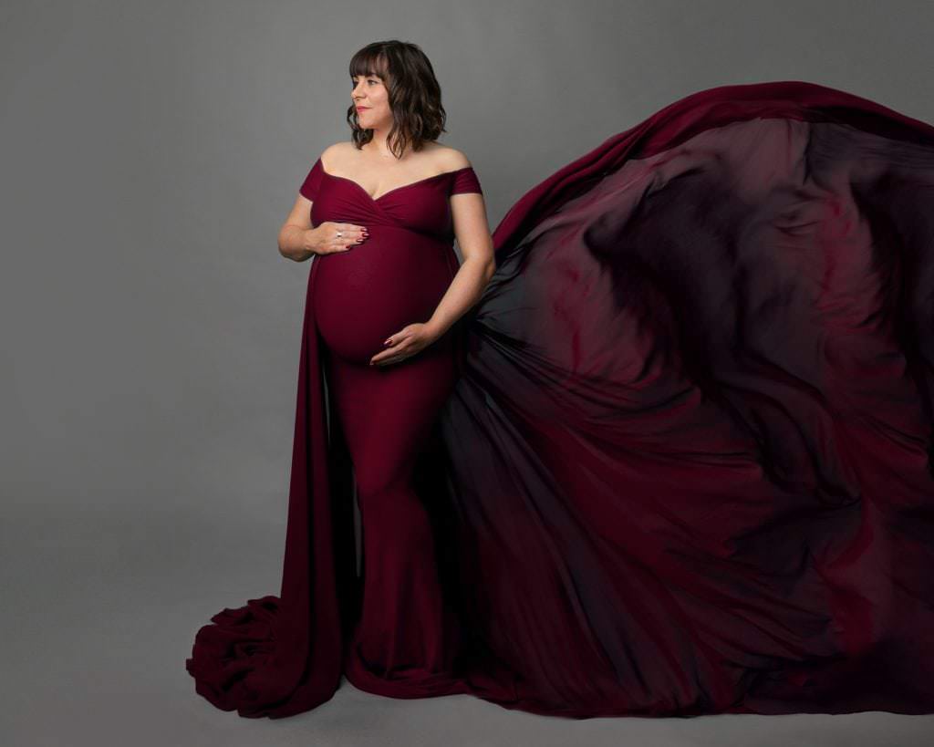 Female wearing a red gown with a high flowing train to the right. She looks left and holds her pregnant bump. Image part of a pregnancy photoshoot in glasgow